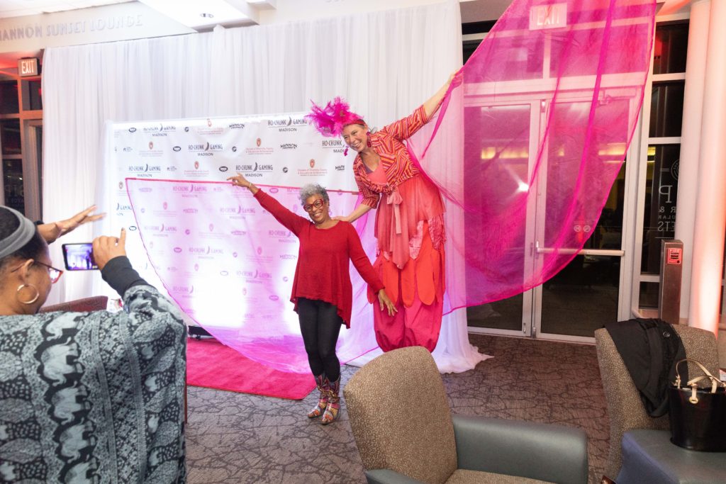 A woman poses for a photo with a stilt walker dressed in a flashy pink outfit with sheer fabric wings on sticks held out to her sides.