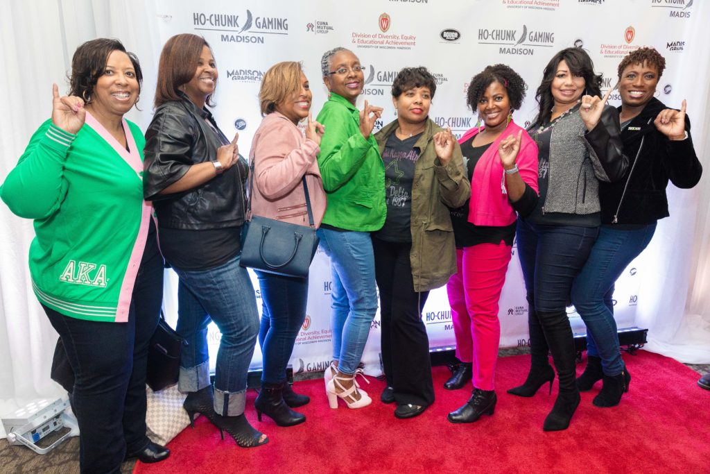 Eight alumni women pose holding up a hand symbol for their sorority, Alpha Kappa Alpha, on a red carpet in front of a backdrop with logos for Ho-Chunk Gaming, CUNA Mutual Group, Rough Sportswear and the DDEEA.