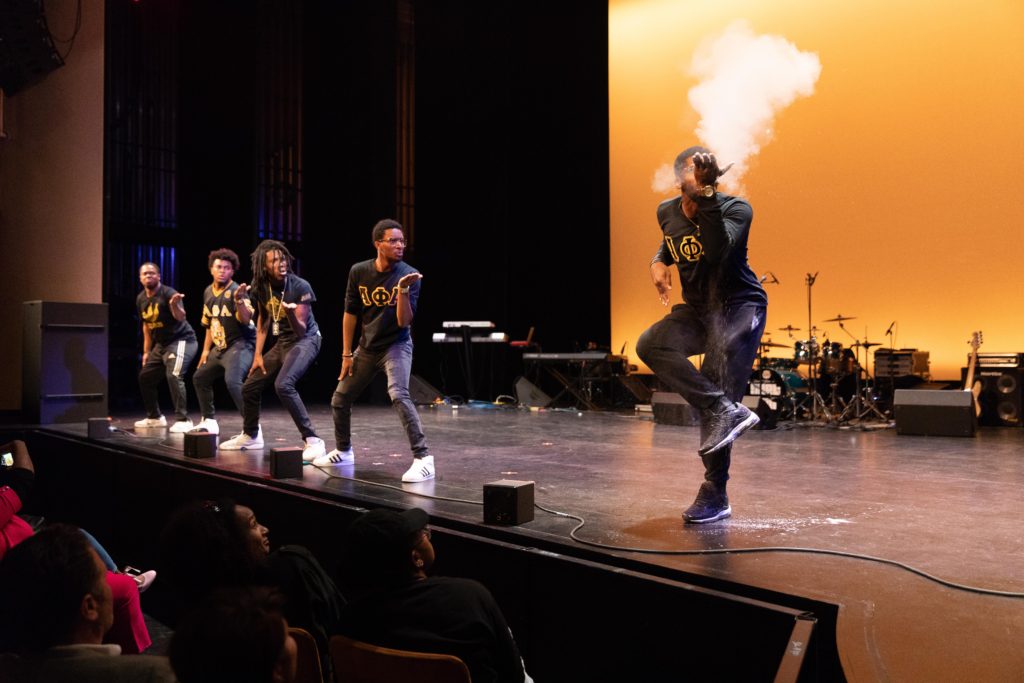 Five members of the Alpha Phi Alpha fraternity stand on stage during a step routine. Four members are looking at the fifth one, who is blowing a white powder into the air while posing standing on one leg.