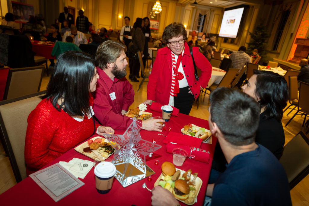 Rebecca blank stands next to a table speaking with four people who are sitting and facing her.