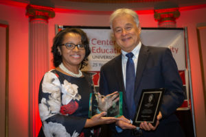 Cheryl Gittens and Walter Lane face the camera smiling while holding two award plaques on stage.