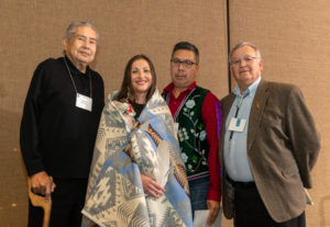 Melissa Metoxen stands with her grandfather and uncle and one more man. Melissa is draped in a blanket with Native designs and is holding a white eagle feather.