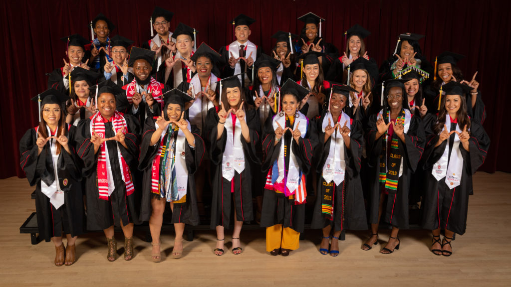 Twenty-six graduating seniors from the PEOPLE program pose in their graduation robes and caps with white stoles draped over their shoulders.