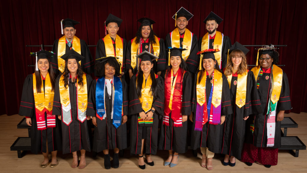 Thirteen graduating seniors from the Posse program pose in their graduation robes and caps with yellow stoles draped over their shoulders.