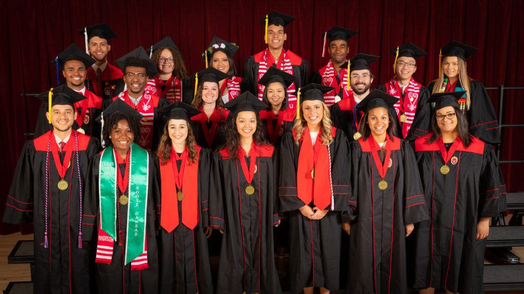 Nineteen graduating seniors from the Powers-Knapp Scholars program pose in their graduation robes and caps with round, gold medallions hanging around their necks