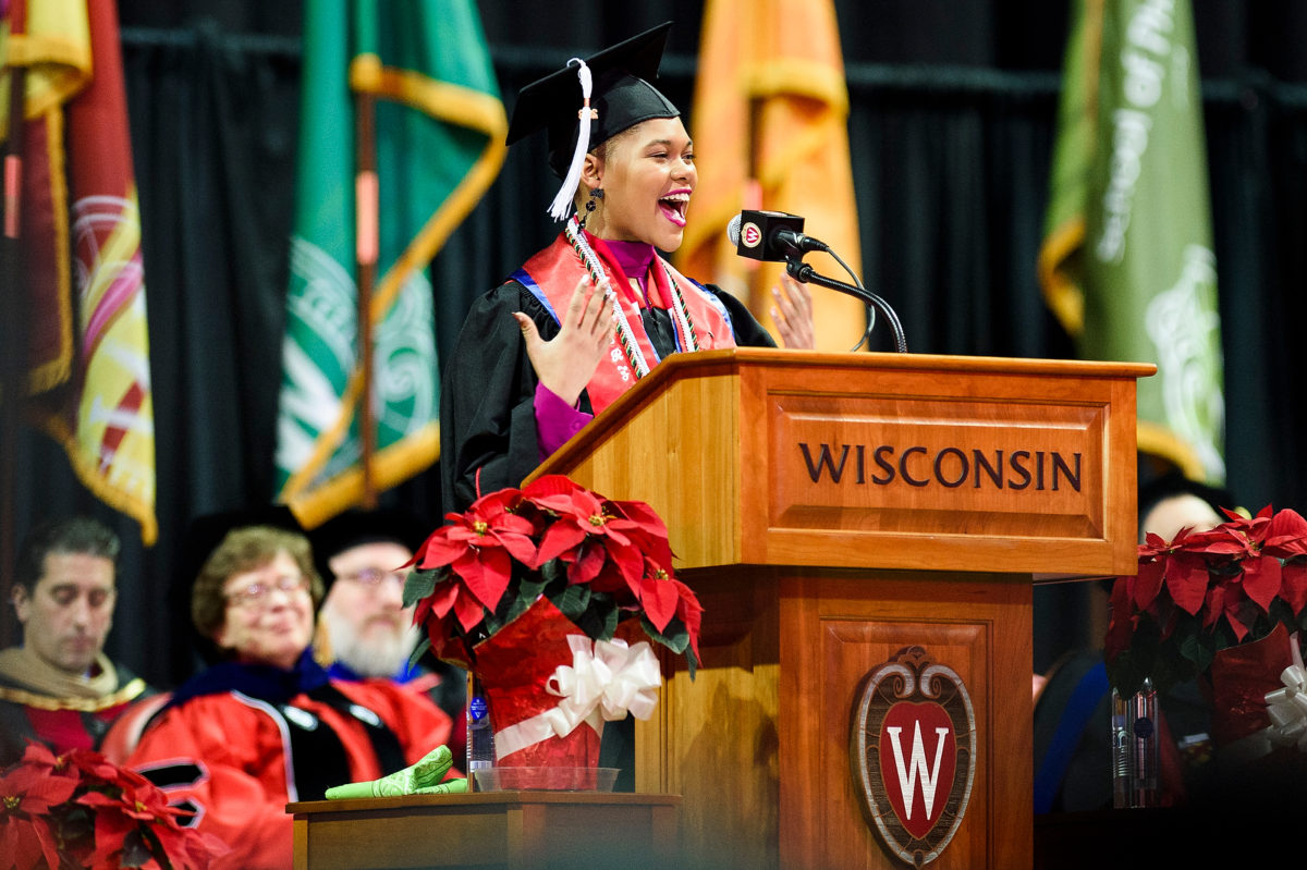 Jamie Dawson stands at a podium bearing the word "Wisconsin" and the UW crest and speaks into a microphone while wearing a traditional cap and gown with colorful stoles. Chancellor Becky Blank and others sit in the background watching her speech.