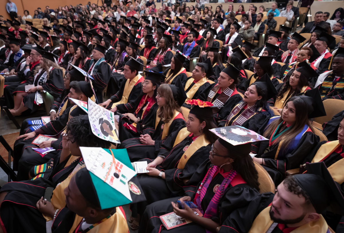 Graduating seniors in caps and gowns sit in auditorium seating in a large room.