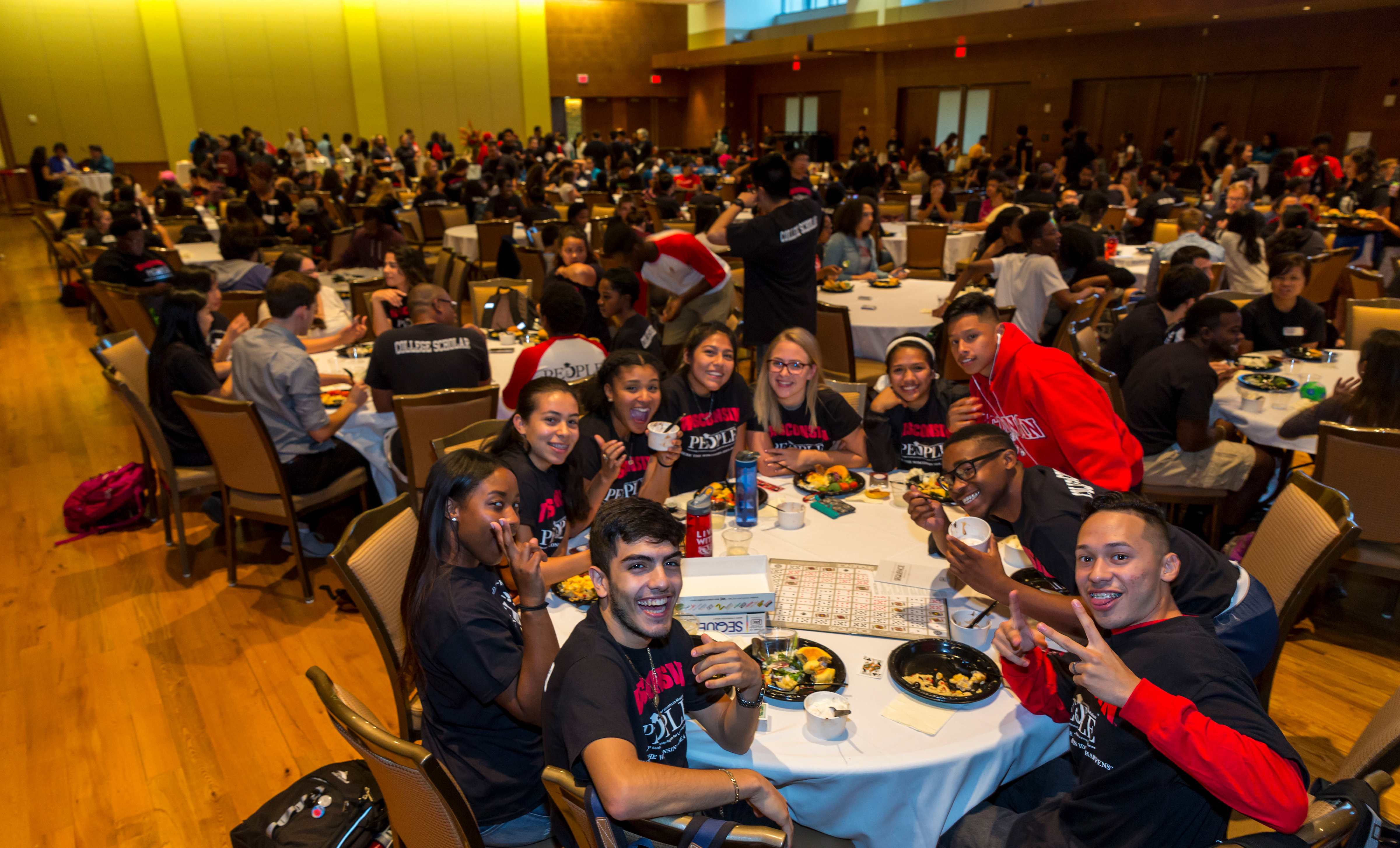 Hundreds of students sit at large, round tables in Union South Varsity Hall during Kickback. In the foreground, a group of students wearing PEOPLE program shirts smile and gesture while looking at the camera.
