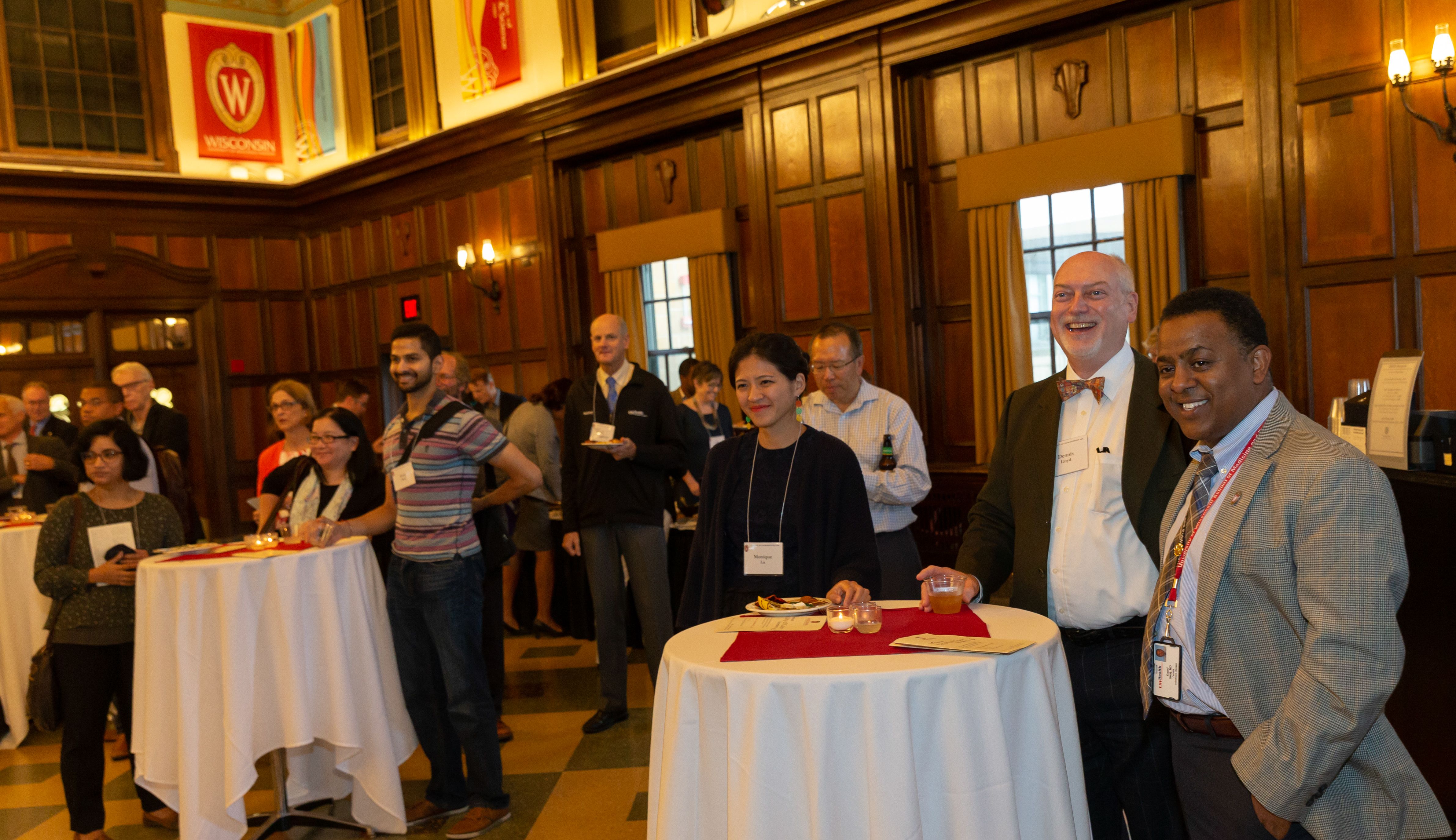 UW–Madison faculty members stand around small tables with drinks while watching a speaker who is not in the picture.