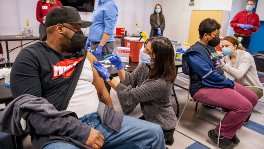 A vaccination clinic with masked people of color receiving vaccine shots from Pharmacy students.