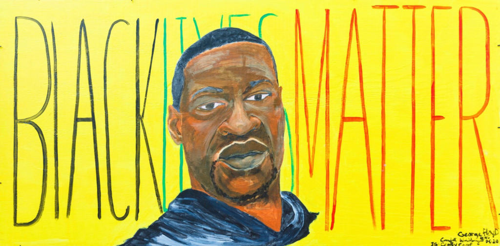 A mural with a yellow background and a painting of George Floyd with the words "Black Lives Matter" written behind his head in black, green and red lettering.