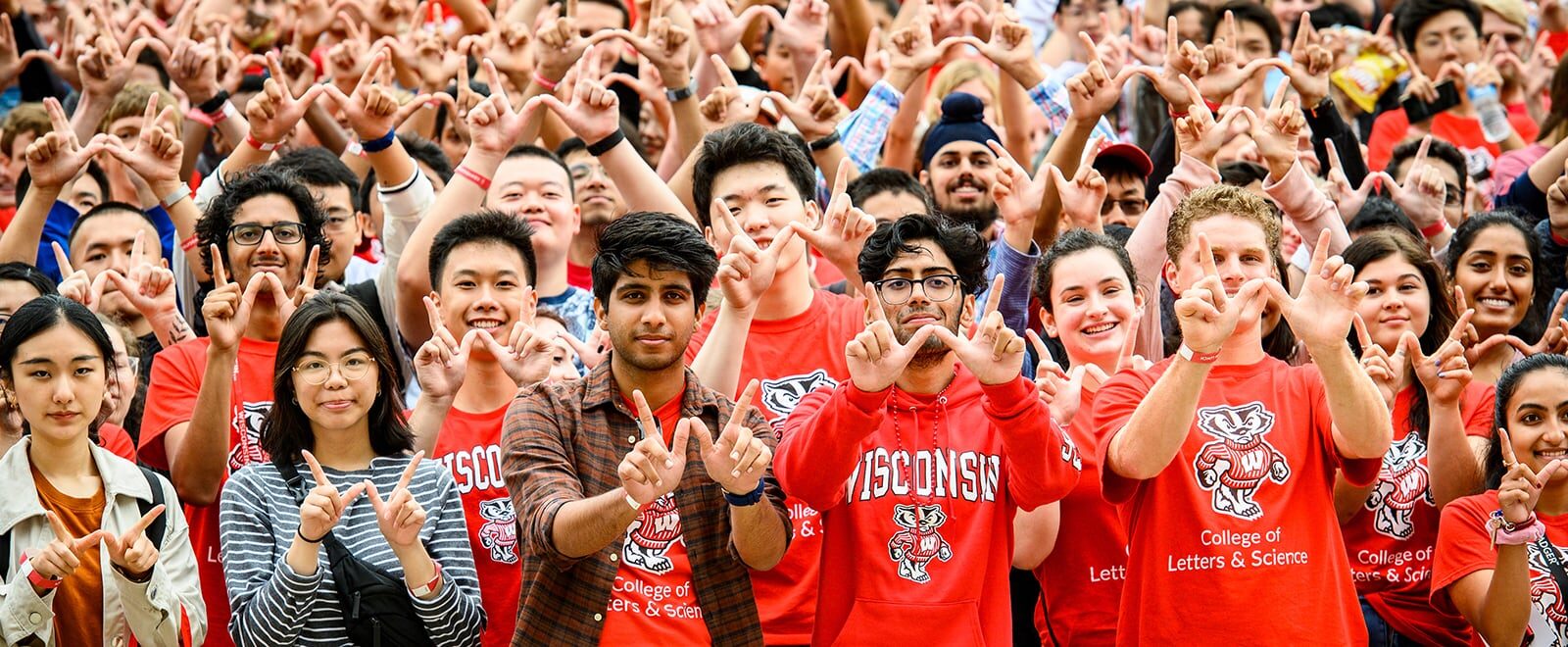 Hundreds of students, many wearing UW–Madison shirts, make the “W-sign” with their hands.