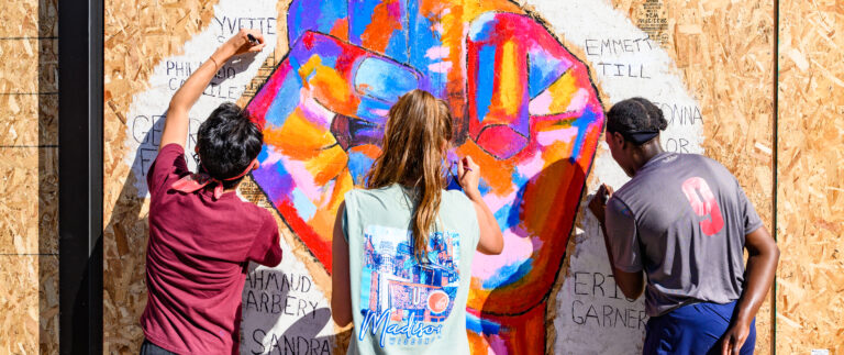 Three students paint a colorful mural of a fist with names around it