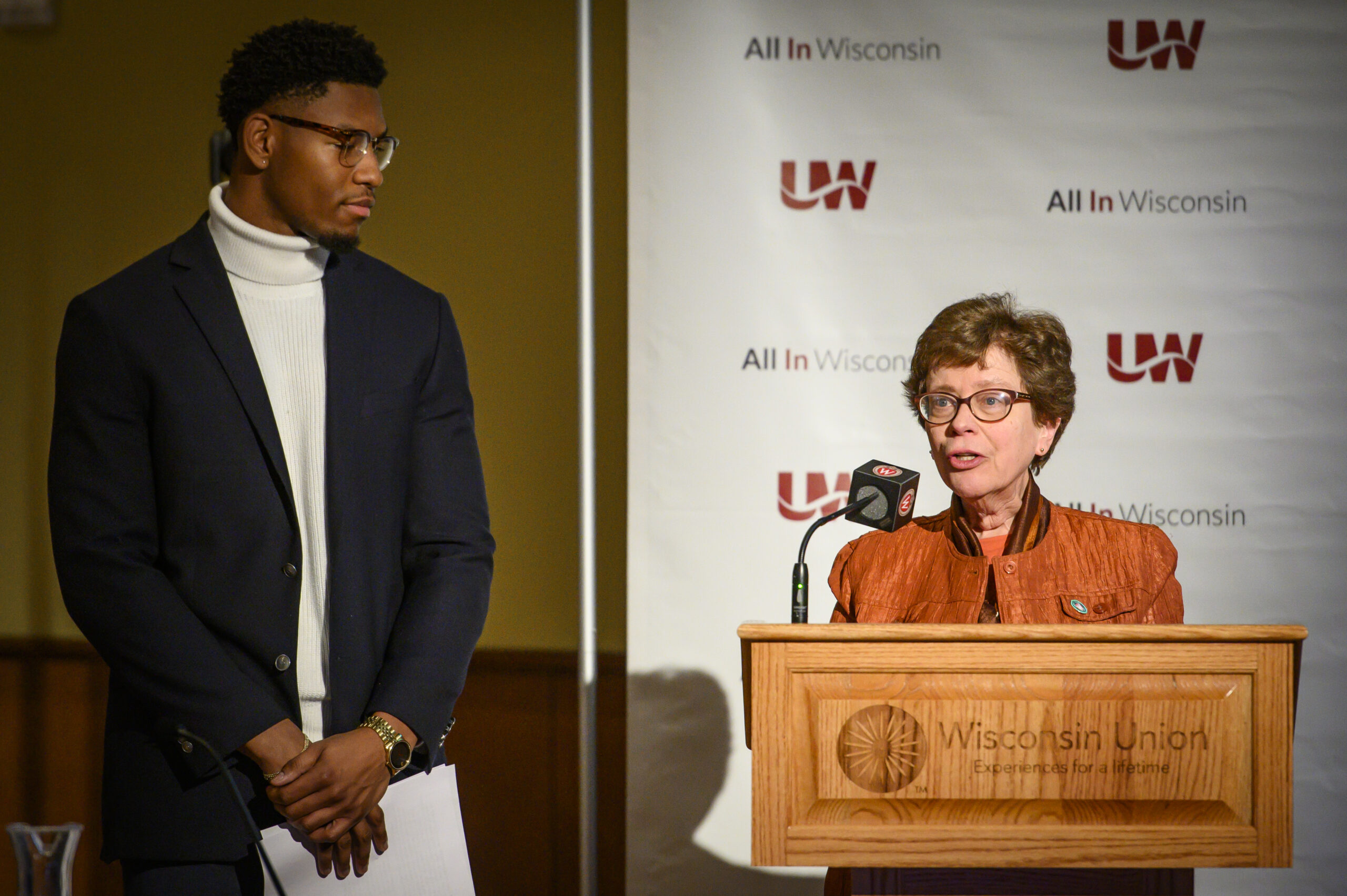 Chancellor Rebecca Blank stands at a podium speaking into a microphone with a student standing next to her.