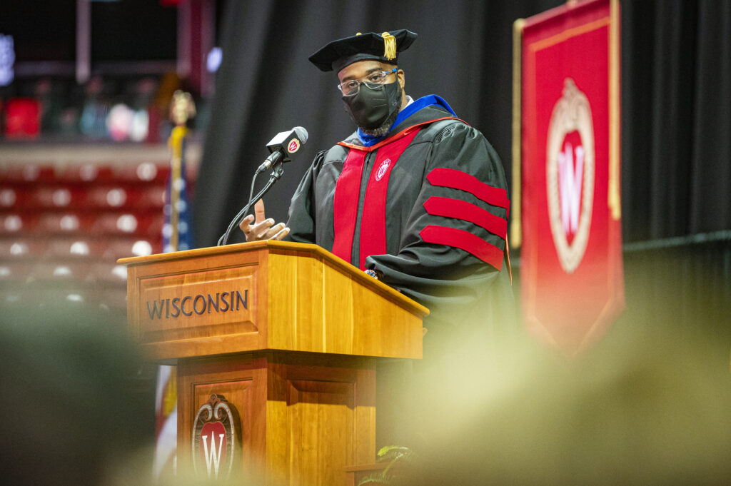 LaVar Charleston speaks into a microphone at a podium while wearing academic regalia and a cloth face mask.