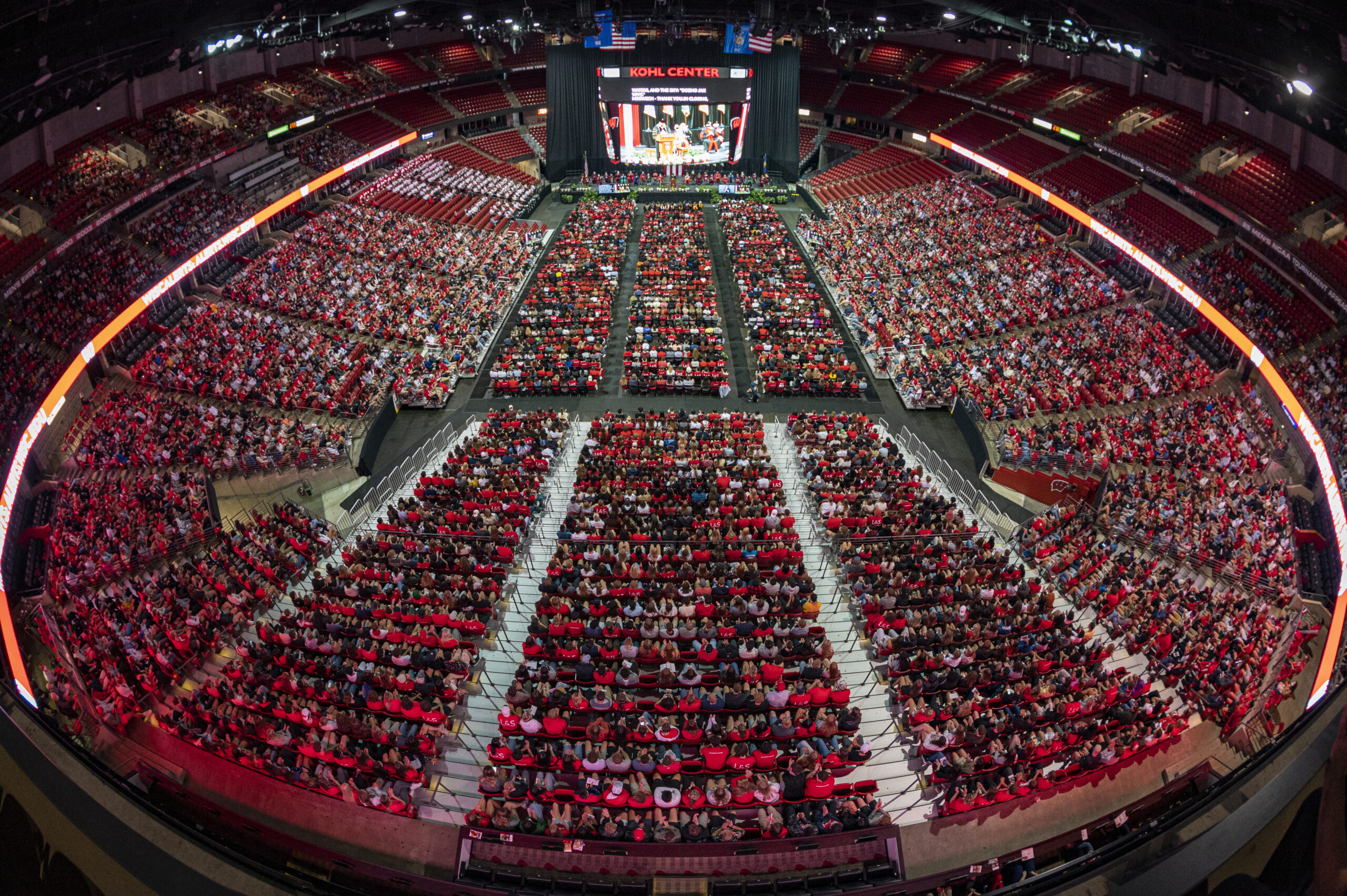 Overhead wide-angle photo of thousands of people sitting in chairs in the Kohl Center facing a stage.
