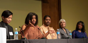Five people sit at a table with microphones during a panel discussion. One of them is speaking and gesturing.
