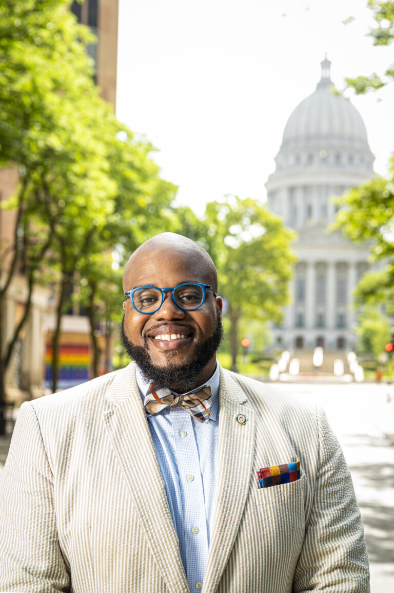 LaVar Charleston wearing an off-white suit, multicolored bow tie and pocket square and blue glasses smiles on State Street with the Wisconsin State Capitol building in the background.