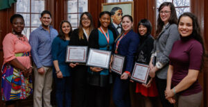 Nine young adults in business attire pose standing for a photo. Four of them in the middle of the group are holding framed certificates and wearing medallions.