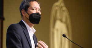 Russell Jeung speaks into a microphone while wearing a black face mask.