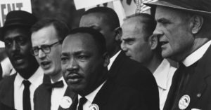 Black-and-white photo of Dr. Martin Luther King Jr. marching with Black and white men holding signs during a protest.