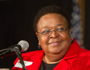 Jackie DeWalt wearing a red suit jacket smiles as she speaks at a microphone.