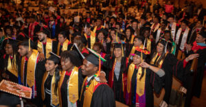 A large group of college graduates in academic attire stand with their arms around each other while smiling and singing in Shannon Hall.