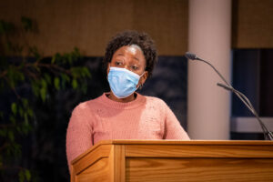 Shenikqua Bouges speaks at a podium wearing a pink sweater and surgical mask.