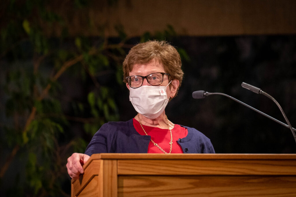 Chancellor Blank speaks at a podium wearing a white face mask, red shirt, purple cardigan and glasses.
