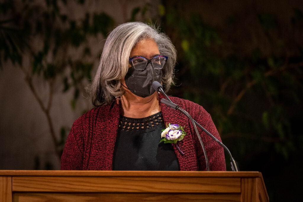Sherl Henderson speaks at a podium wearing a black blouse and maroon cardigan.