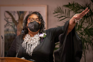 Carola Peterson-Gaines gestures while speaking at a podium weaering a black dress and large silver and white necklace.