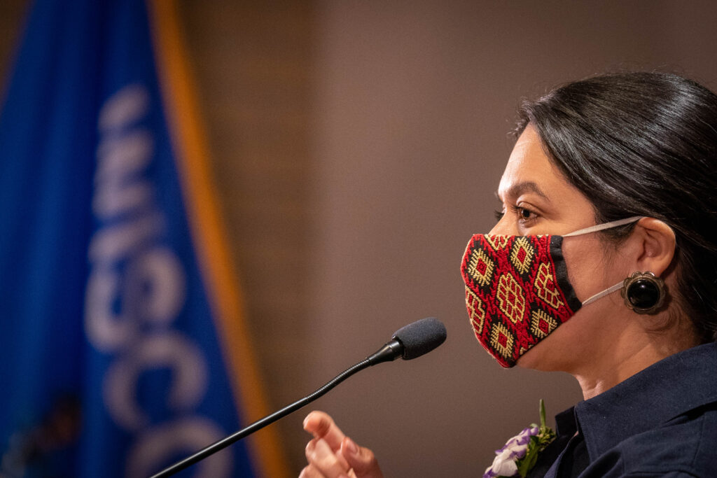 Carolina Sarmiento speaks into a microphone wearing a colorful, embroidered cloth face mask.