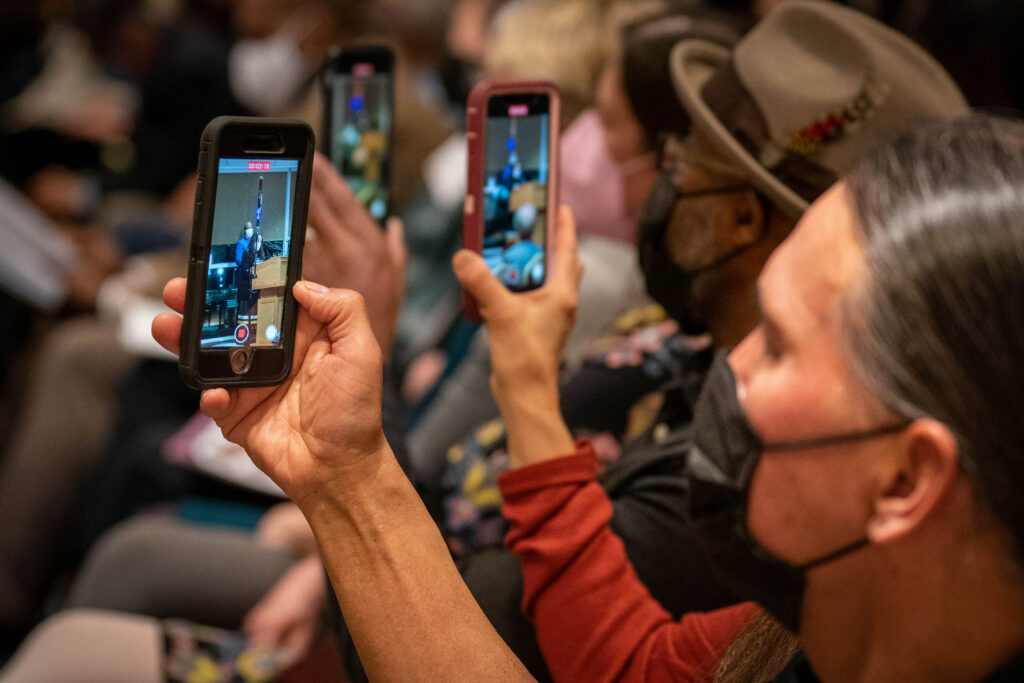 Audience members in face masks hold up their phones to record the presentations.