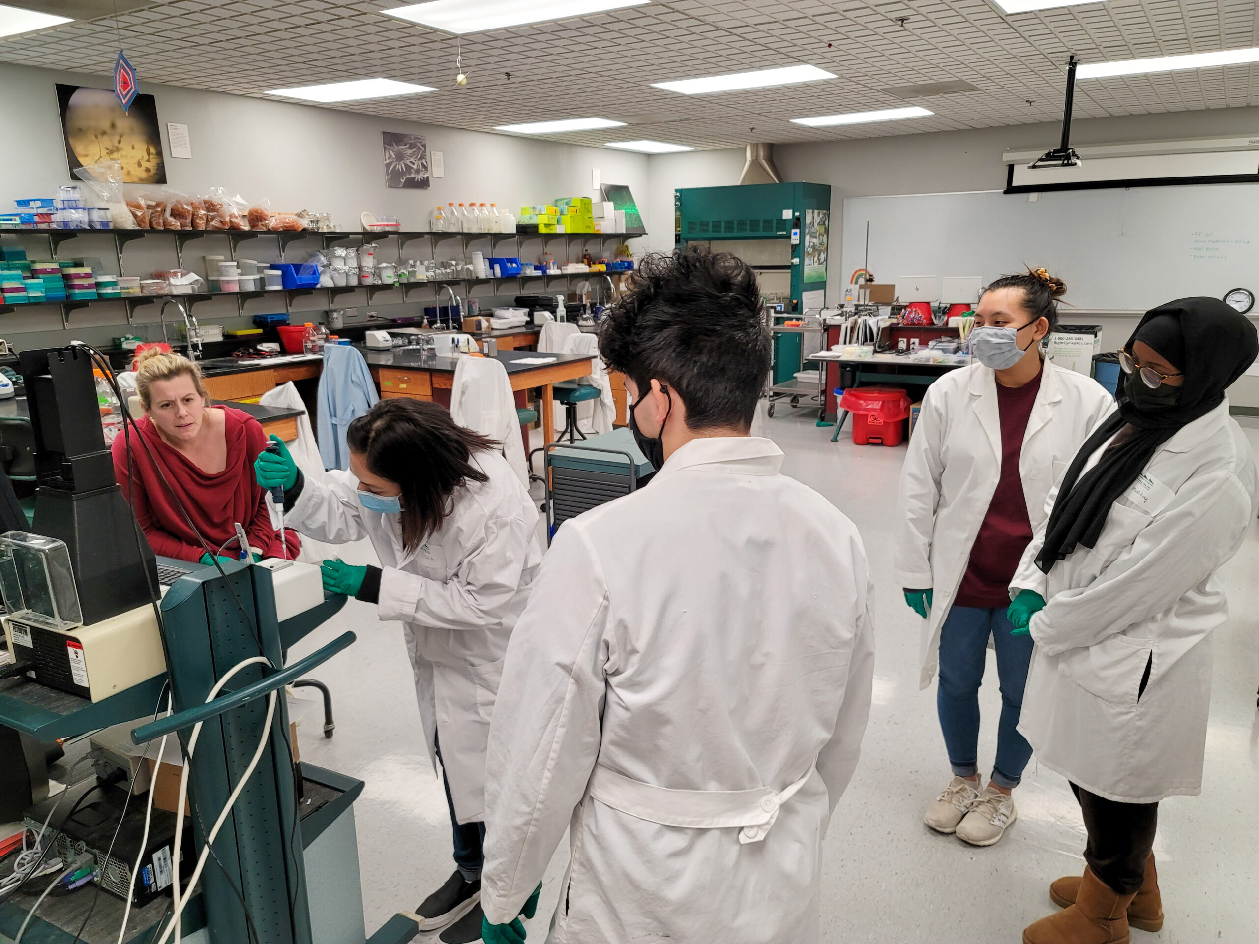 Four students work in a science lab wearing white lab coats, green surgical gloves, and face masks while an instructor in a red shirt watches.
