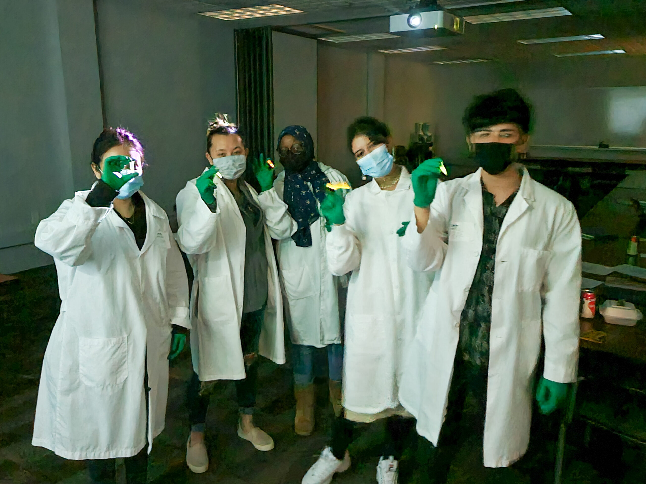 Five college students in white lab coats stand holding small white things in a dark room.