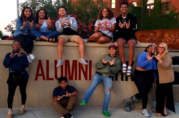 A group of college students make a W sign with their hands while standing and sitting around the Alumni Park sign near the Red Gym.