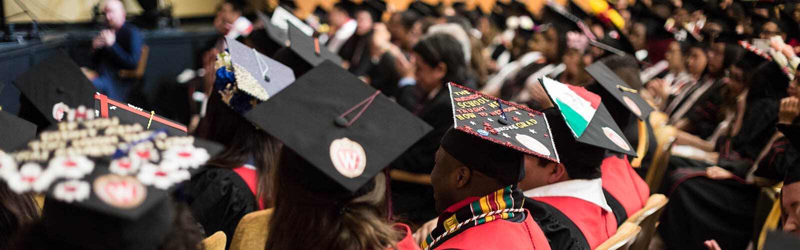 College graduates wearing decorated graduation caps sit in a theater at a graduation ceremony.