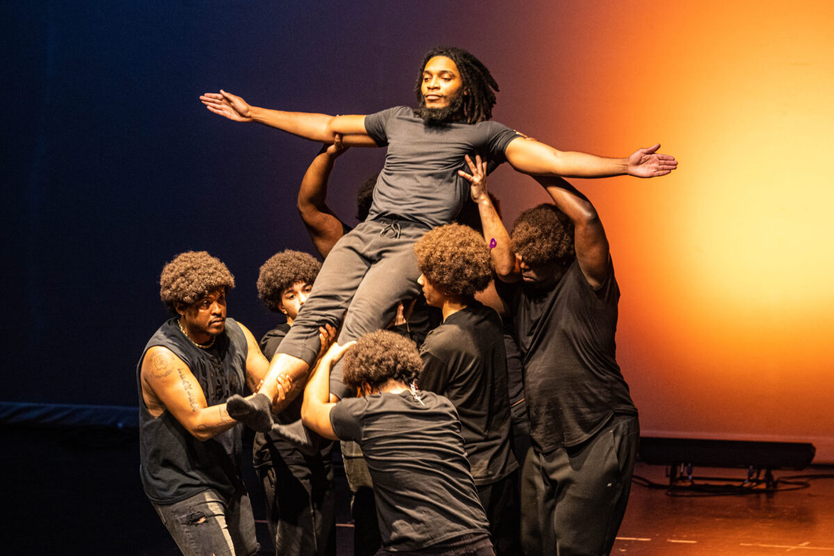 Performers dressed in black on stage catch one person as they fall backward with arms spread wide.