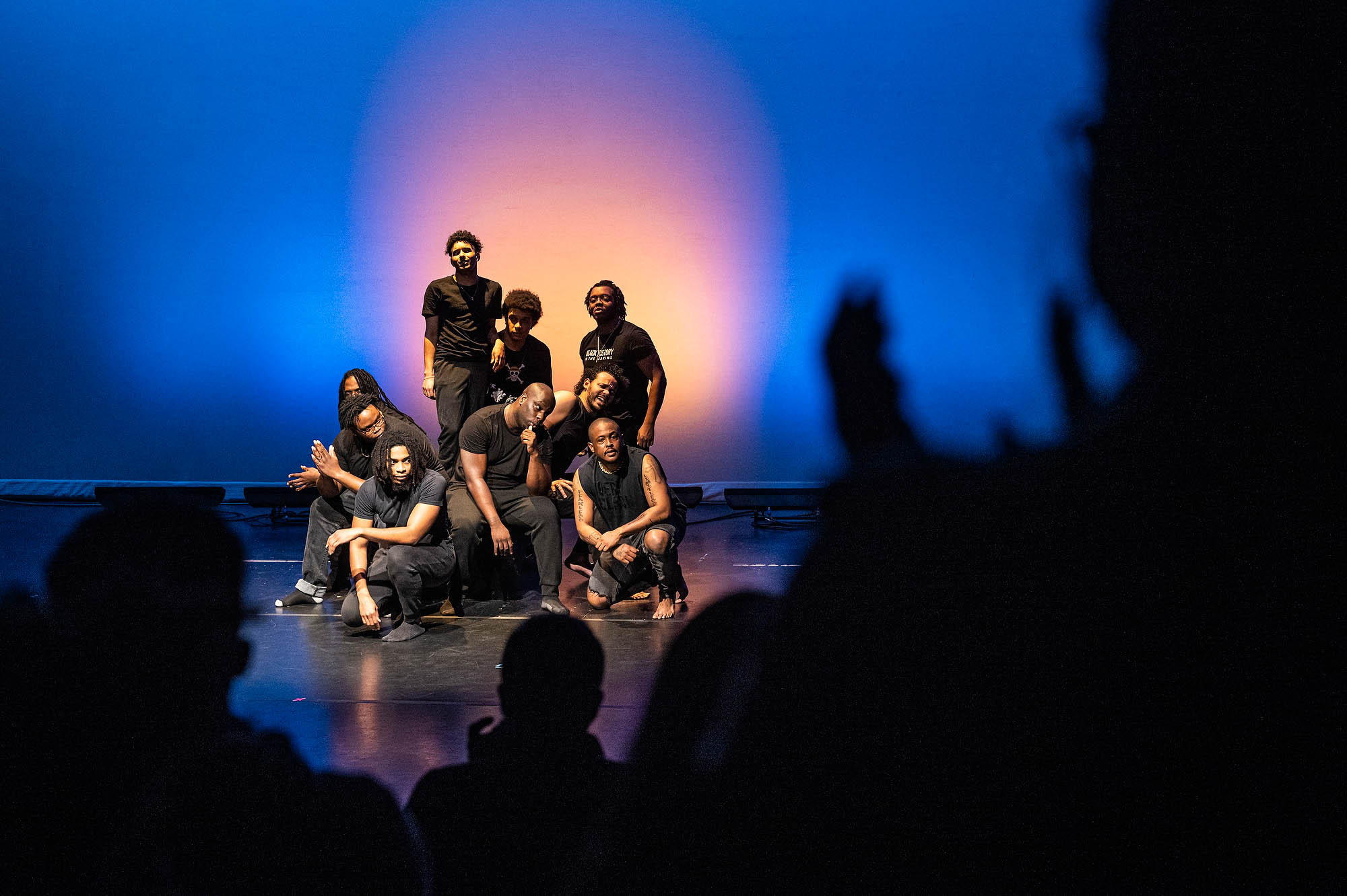 A group of performers in black pose in a group while recieving a standing ovation from an audience.