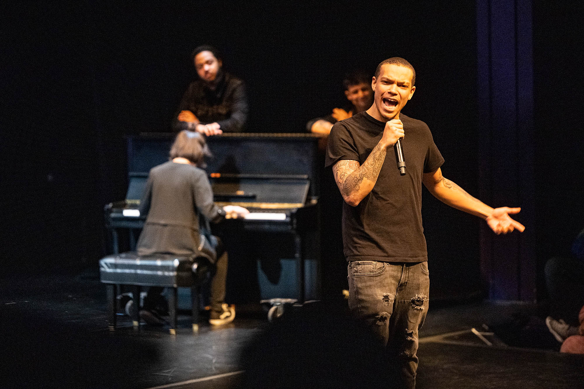 A performer dressed in black speaks into a microphone while behind him other performers dressed in black play a piano and watch.