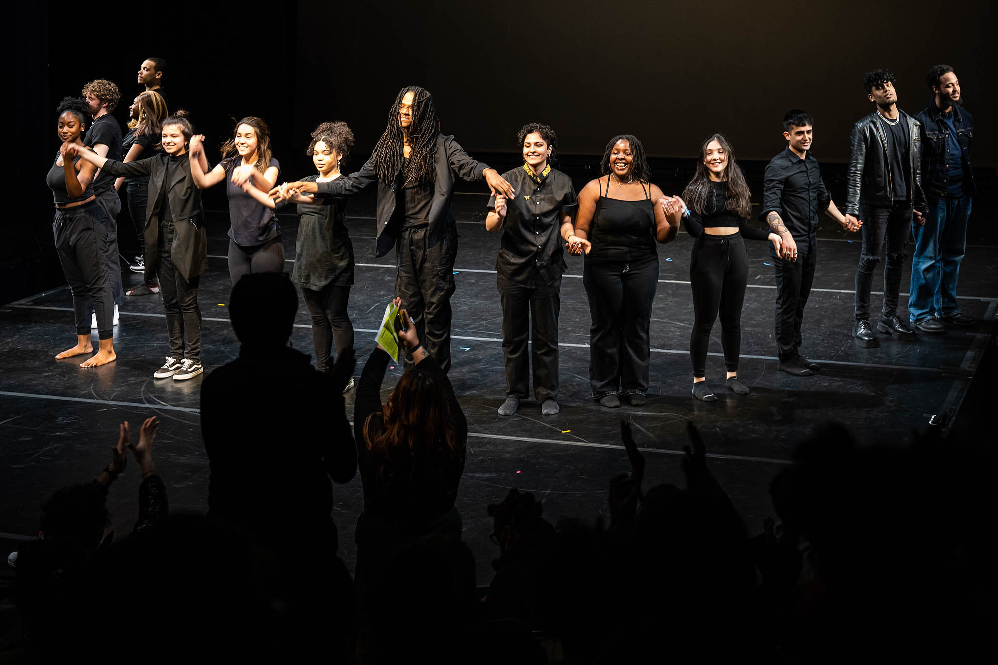 A large group of performers wearing black hold hands and bow on stage.