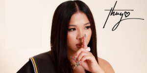 The singer Thuy holds her index finger in front of her mouth and nose. Over her shoulder is a stylized "thuy" signature with a heart symbol.