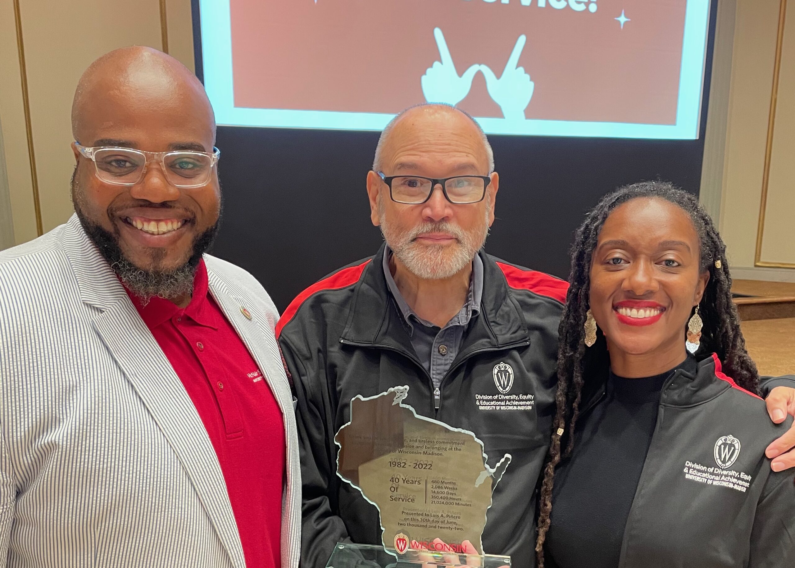 Three people stand shoulder to shoulder and pose for a photo. The center figure holds a glass award in the shape of the state of Wisconsin.