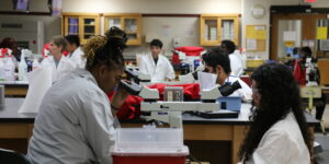 High school students in white lab coats use scientific equipment in a lab.
