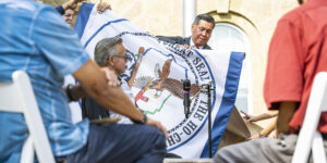 A group of men hit a large drum in the foreground, while behind them a man in a suit holds the flag of the Ho-Chunk Nation.