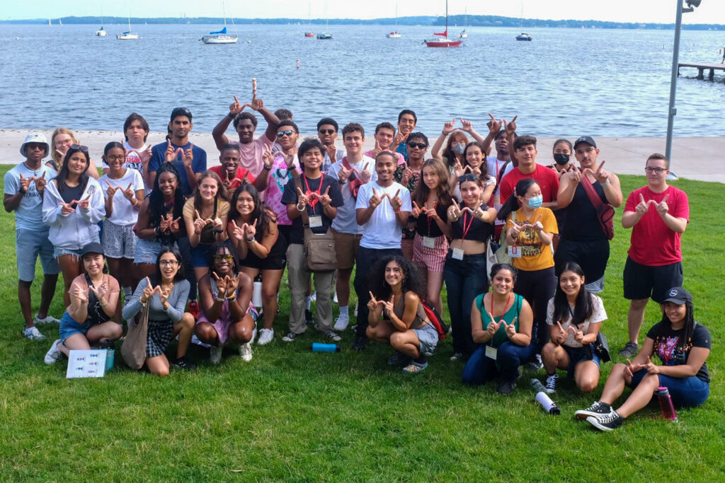 A large group of college students pose for a group photo making W signs with their hands on a grassy area in front of Lake Mendota.