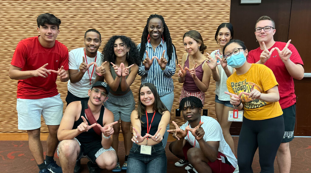 A group of 11 students with a wide range of skin colors and gender expressions make W signs with thier hands and smile for a group photo.