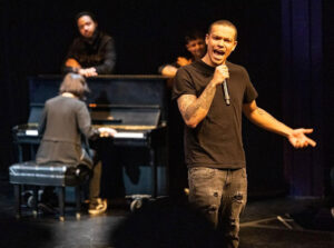 A man with arm tatoos and buzz-cut hair wearing a black t-shirt speakes into a wireless microphone on a stage with musical performers behind him.