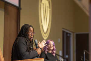 Tiya Miles holds a microphone and gesticulates, with a W crest representing the University of Wisconsin in the background.