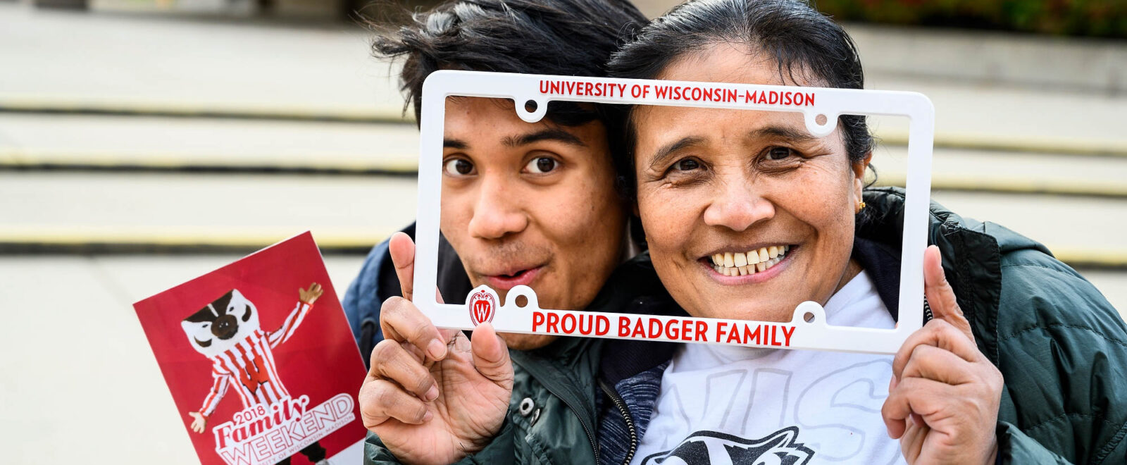 A college student and his mom pose laughing with faces close together framed by a University of Wisconsin license plate frame.